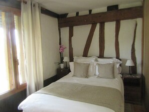 Ein Schlafzimmer mit einem großen Bett, Holzbalken an der Decke, einem Nachttisch mit Lampe und einem Fenster mit Vorhängen.