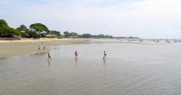 A beach with people walking in shallow water, a sandy shore, and boats in the distance. - Vrbo Property (Andernos-les-Bains)