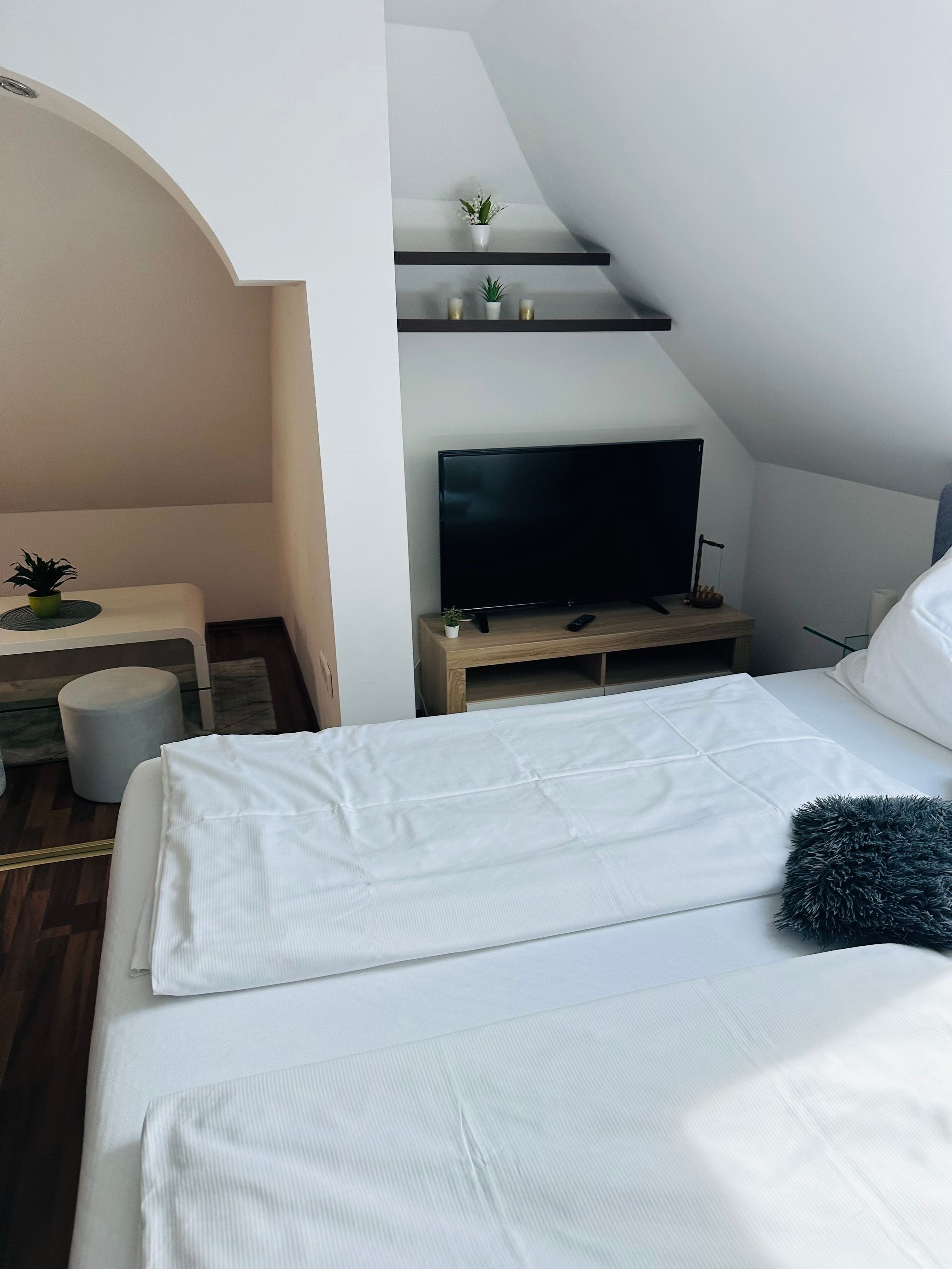 A modern bedroom with a flat-screen TV, a bed with white linens, a small table with a plant, and a staircase with a shelf above.