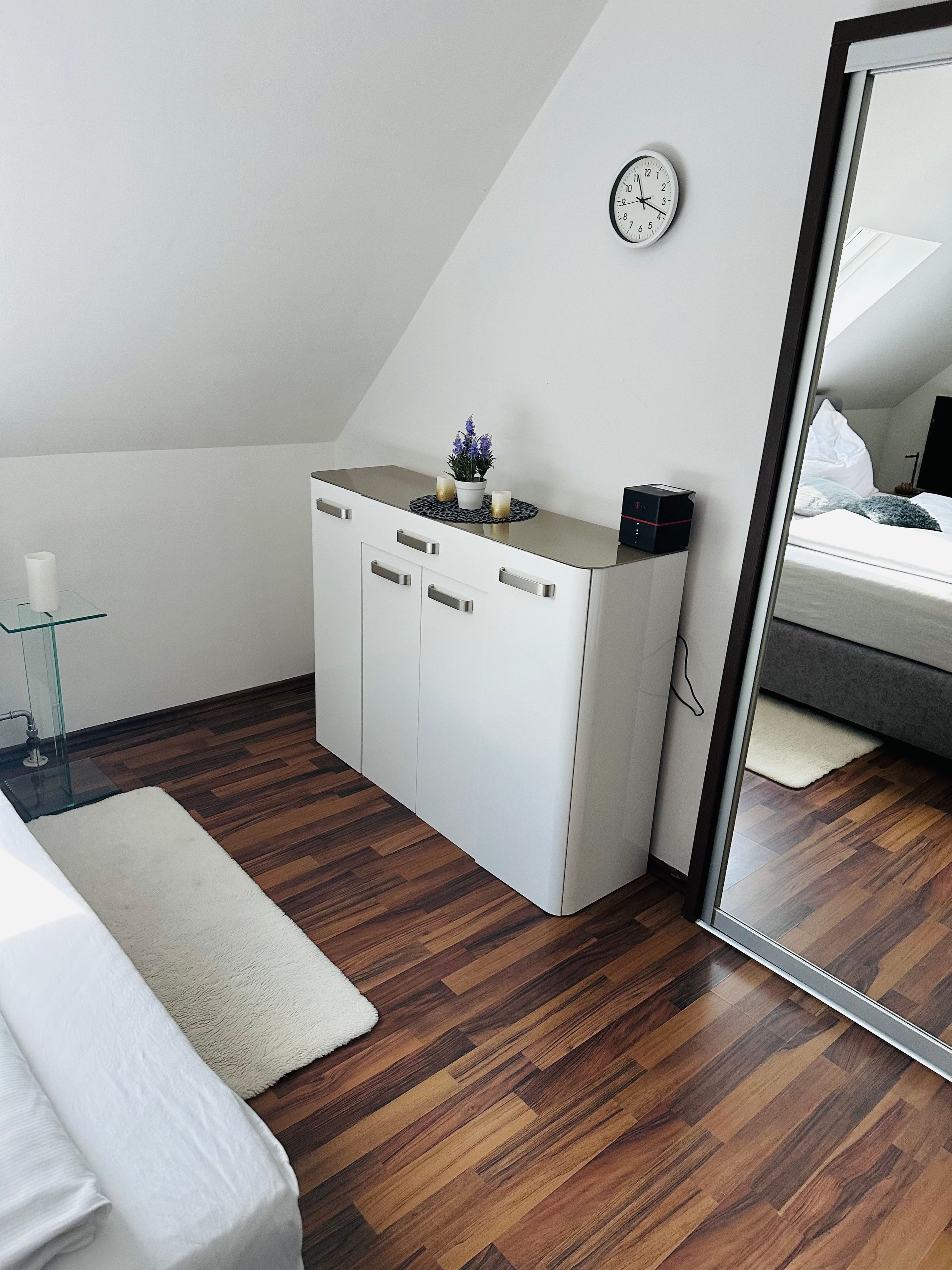 A modern, minimalistic room with a white wardrobe, a small bedside table, a clock on the wall, and a mirror reflecting part of the room.