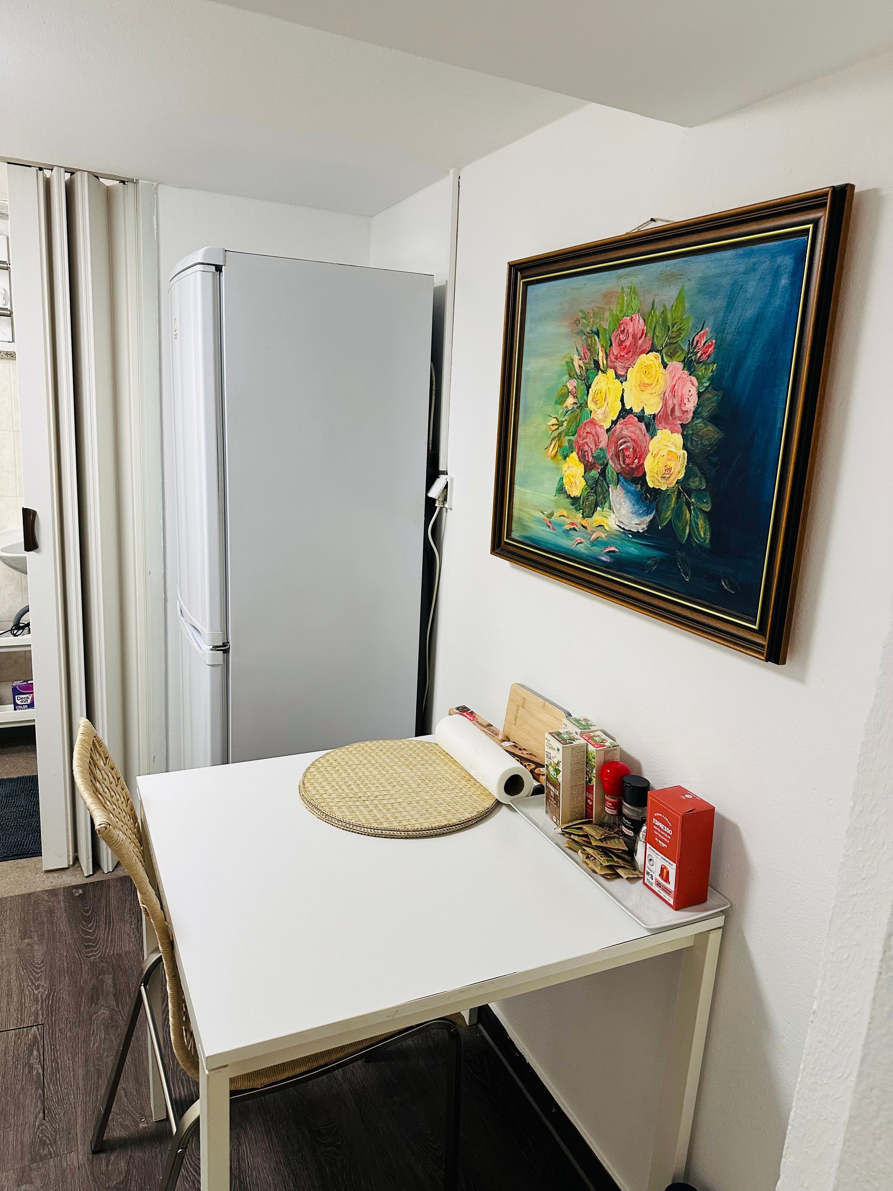 Una habitació petita amb una taula blanca, un quadre amb flors, un refrigerador blanc i una cadira.