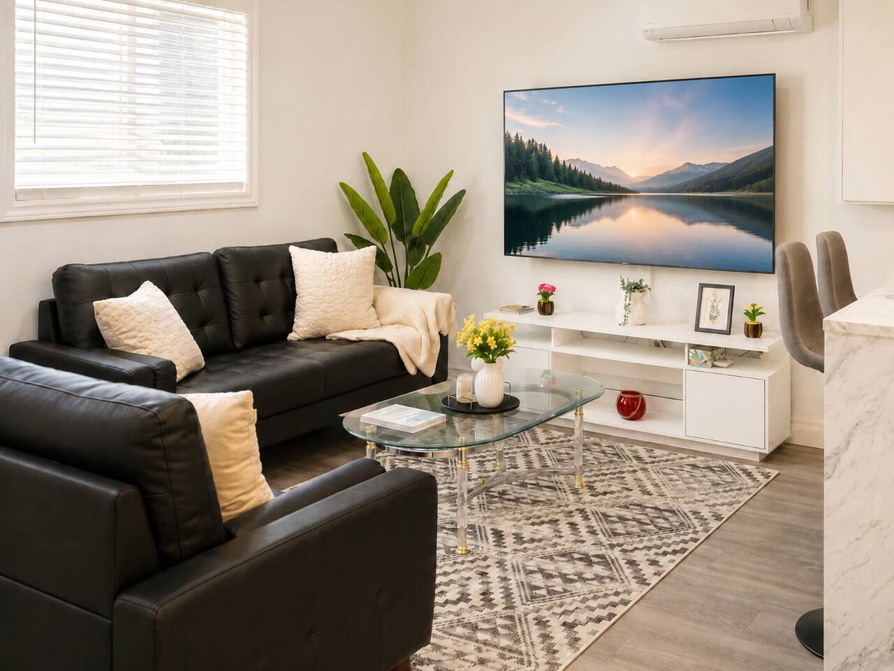 Un salon moderne avec un canapé en cuir noir, une table basse en verre, une console blanche agrémentée d’objets décoratifs et un grand téléviseur à écran plat diffusant une image d’un lac de montagne.