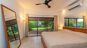 Ein Schlafzimmer mit Bett, Deckenventilator, Klimaanlage und Blick auf Grün durch das Fenster.