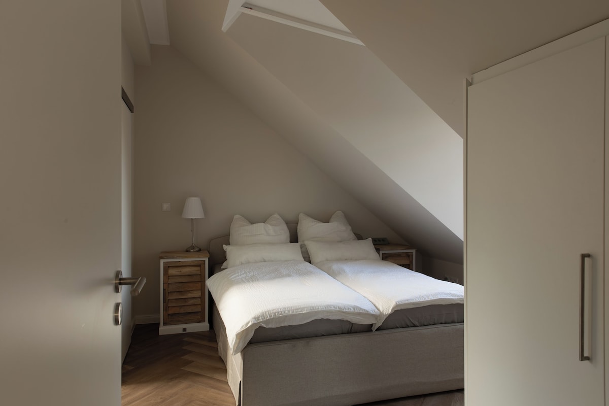 Une chambre avec un plafond en pente, un lit avec une literie blanche, une table de chevet avec une lampe et une commode en bois.