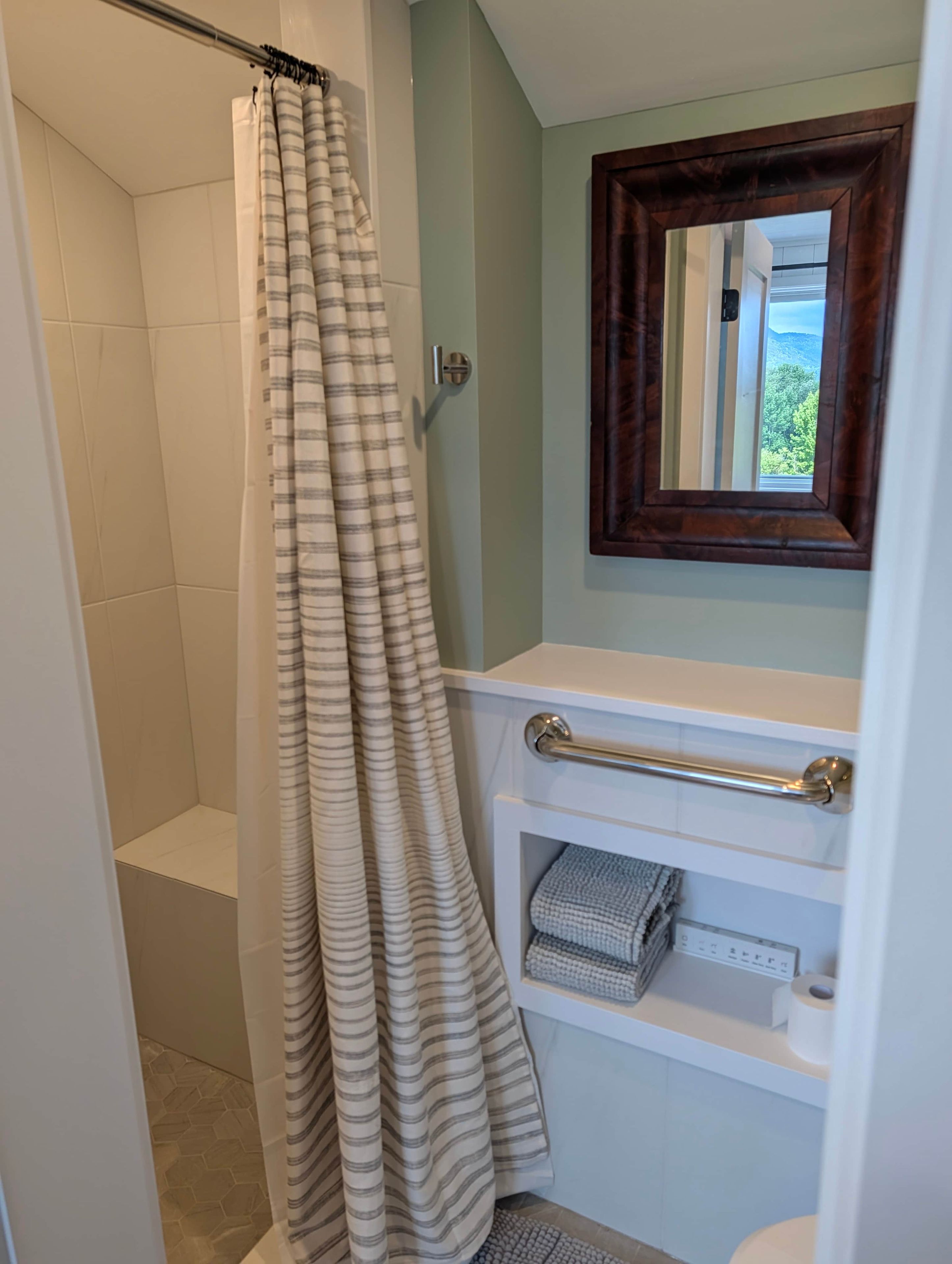 A bathroom with a shower curtain, a towel rack, a toilet paper holder, and a window with a view of greenery.