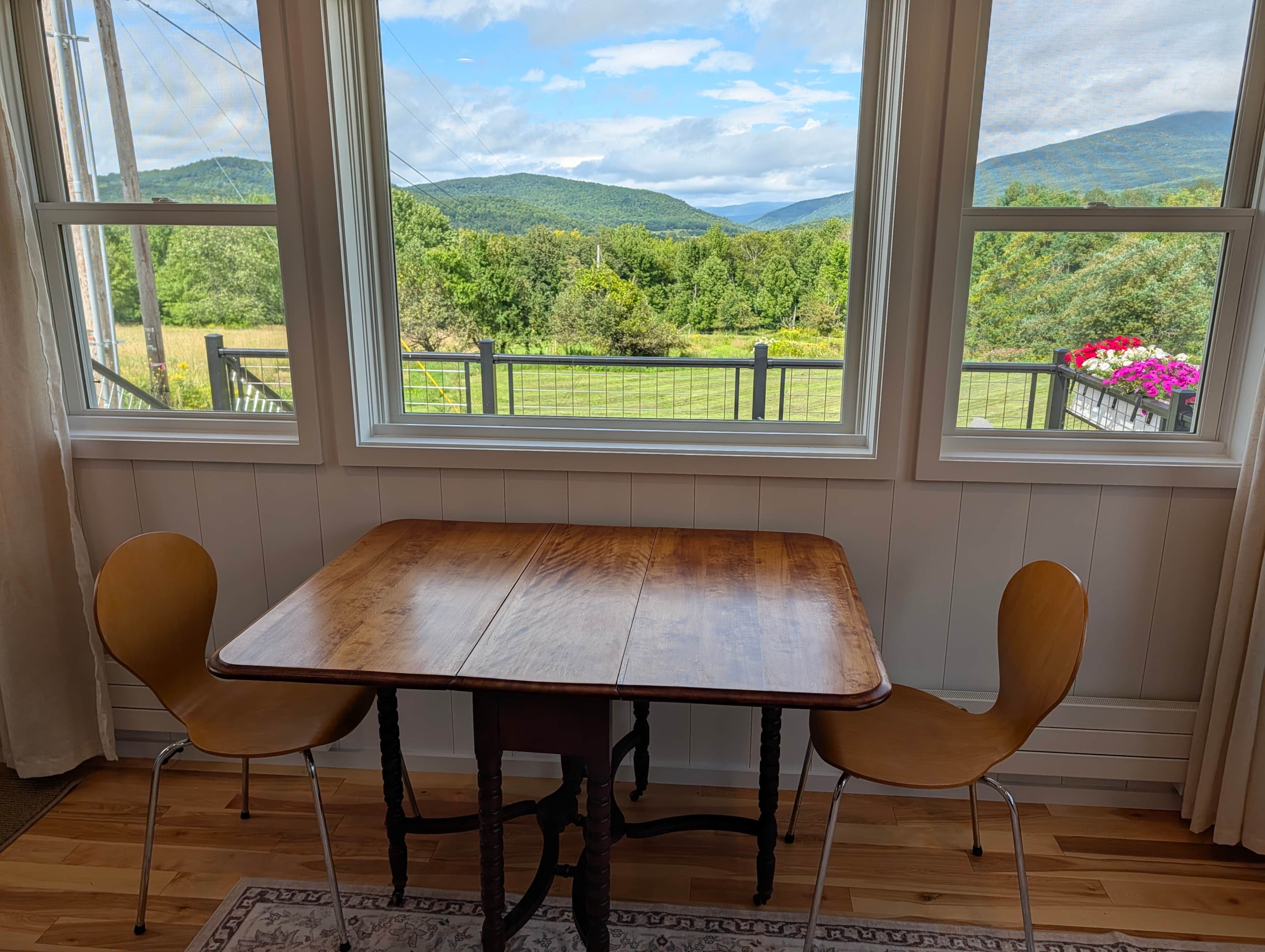 Une pièce avec une table en bois et deux chaises, de grandes fenêtres donnant sur des collines verdoyantes.