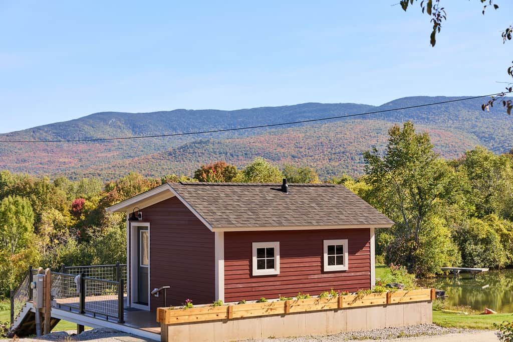 A small red cabin with a door and windows, a wooden deck, and a view of a lake and mountains in the background.