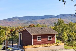 Une petite cabane rouge avec une porte et des fenêtres, une terrasse en bois et une vue sur un lac et des montagnes en arrière-plan.