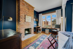 Une pièce avec une cheminée en briques, un bureau, une chaise, une fenêtre avec des rideaux et une vue sur les bâtiments à l’extérieur.