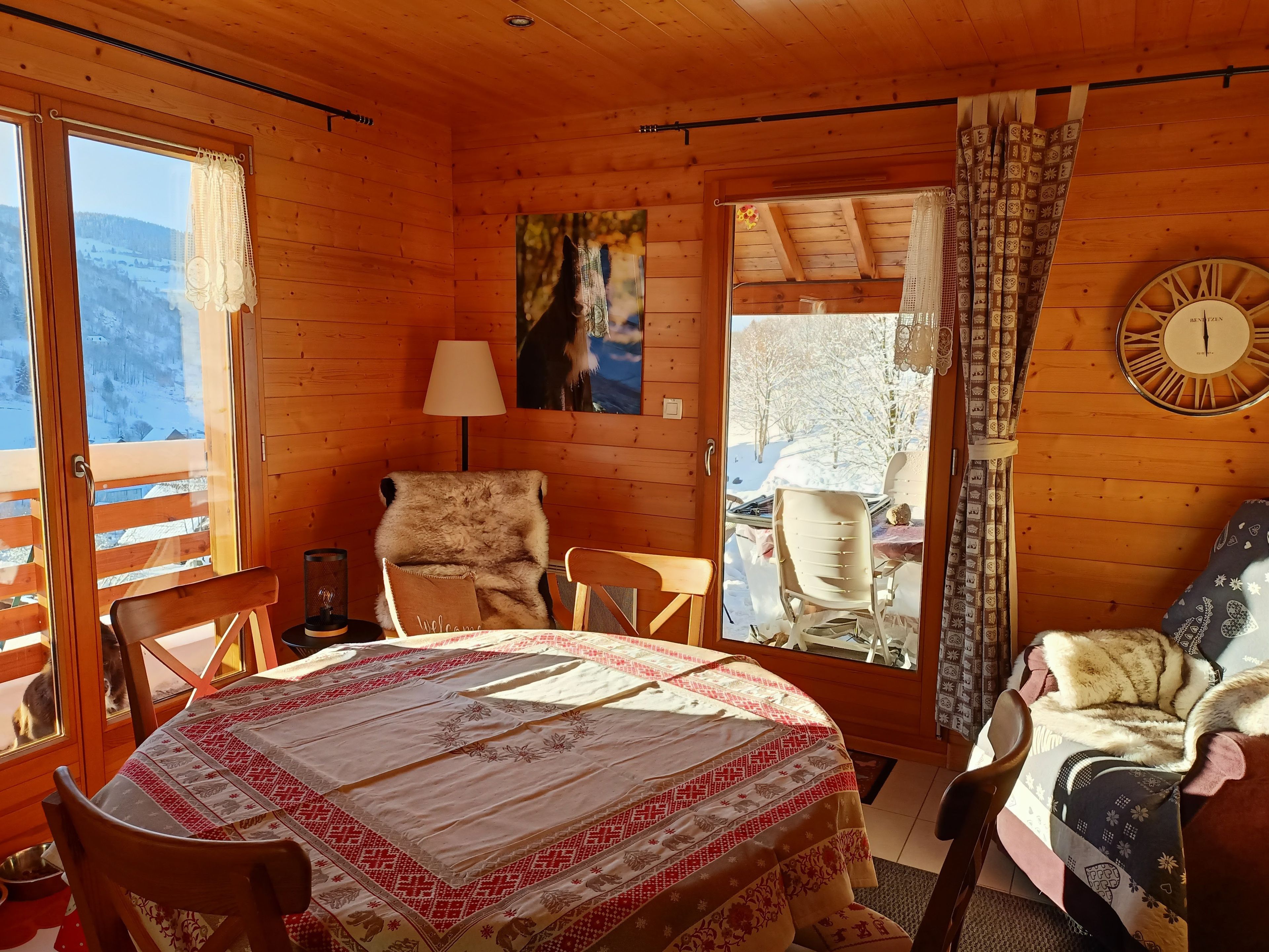 L’intérieur chaleureux d’un chalet, avec un décor en bois, une table à manger entourée de chaises, un canapé, une horloge et une fenêtre donnant sur des montagnes enneigées.