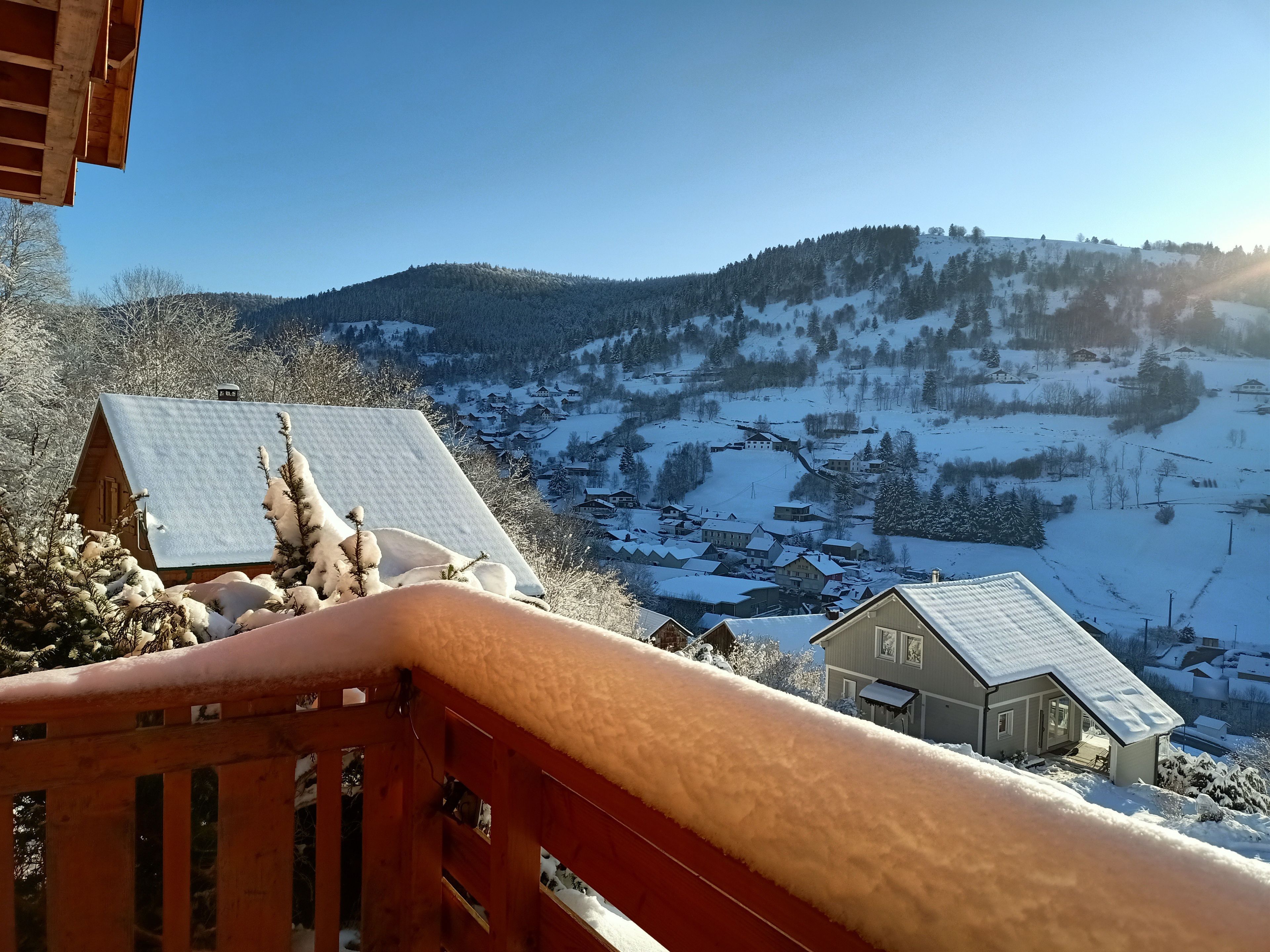 Een houten chalet met een met sneeuw bedekte dak, een rode balustrade en uitzicht op een besneeuwd dorp en bergen op de achtergrond.