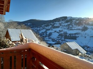 Une cabane en bois, dont le toit est recouvert de neige, avec une balustrade rouge, et qui offre une vue sur un village enneigé et des montagnes enneigées en arrière-plan.