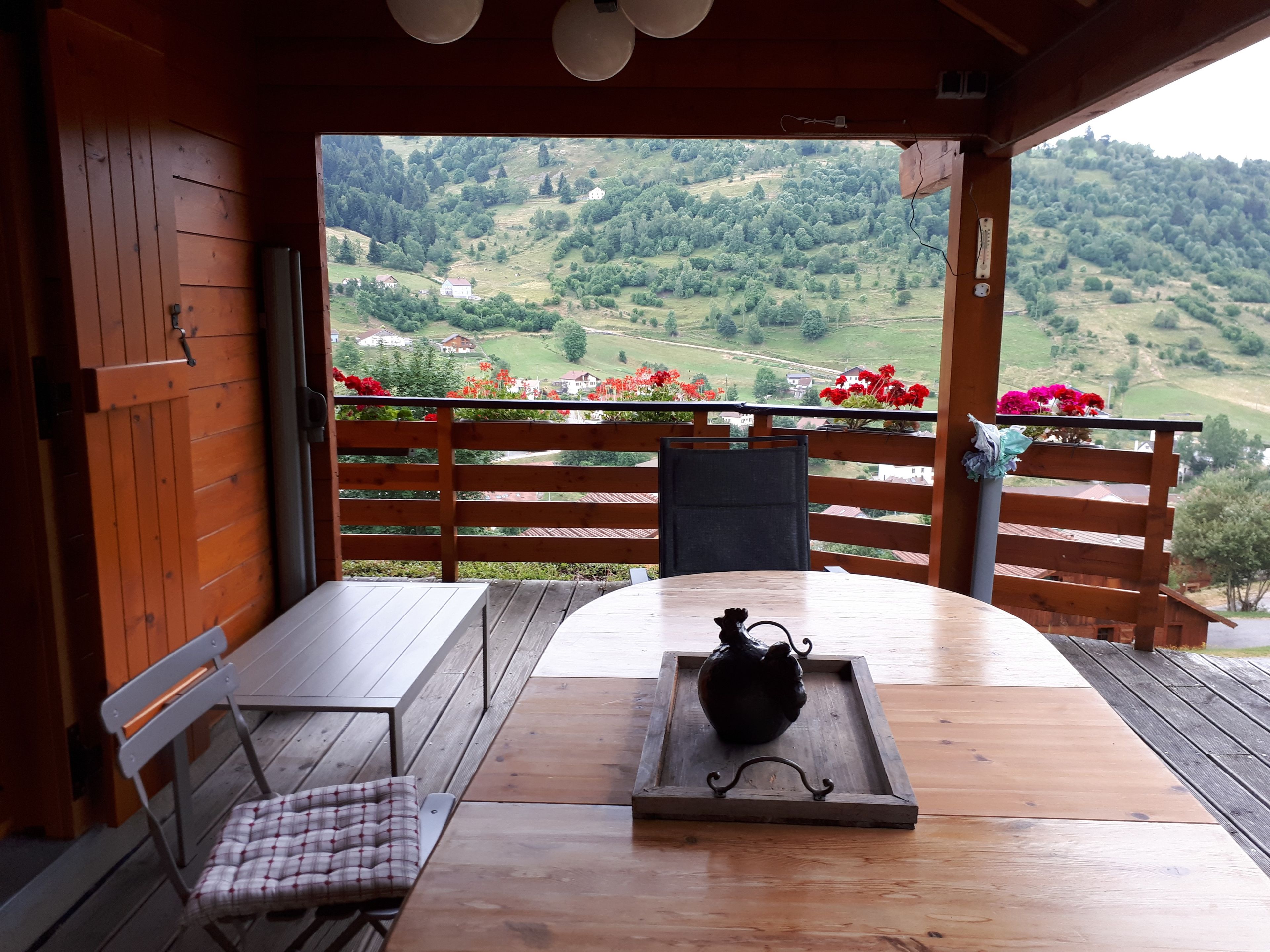 Una terraza de madera con una mesa y sillas, un florero decorativo, y vistas a un valle verde con casas.