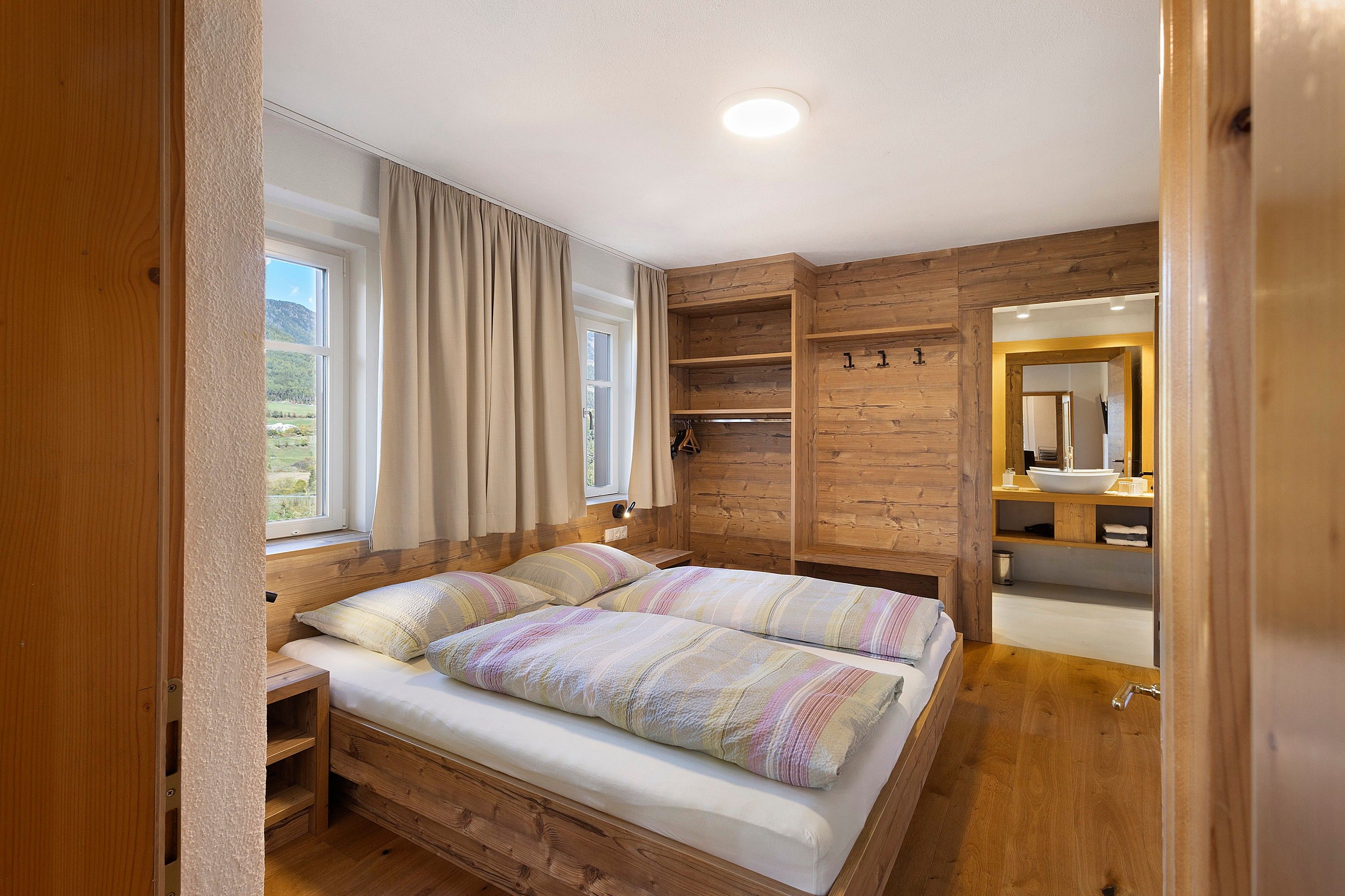 Une chambre avec un lit en bois, une fenêtre avec des rideaux, une étagère intégrée et une salle de bain avec un miroir et un lavabo.