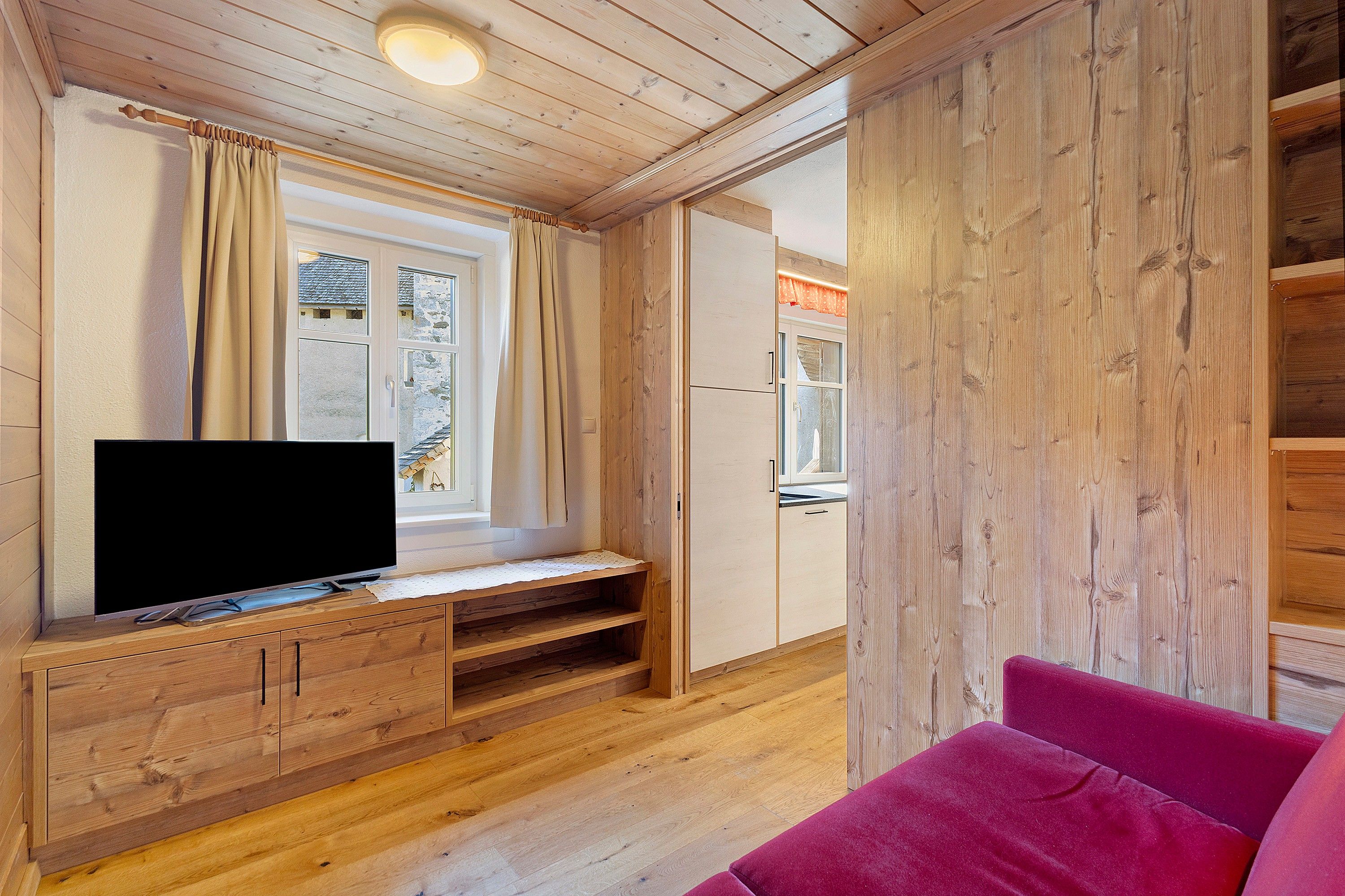 Un intérieur en bois avec un téléviseur posé sur un meuble en bois, une fenêtre avec des rideaux et un canapé rouge.