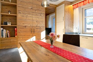 Une table à manger en bois, agrémentée d’un chemin de table rouge, d’un vase avec des fleurs et d’une bibliothèque contenant des livres et une radio.
