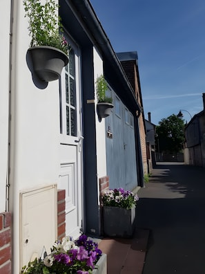 Eine enge Straße mit einem weißen Gebäude, das eine Tür, Fenster, Blumentöpfe und einen klaren Himmel zeigt.