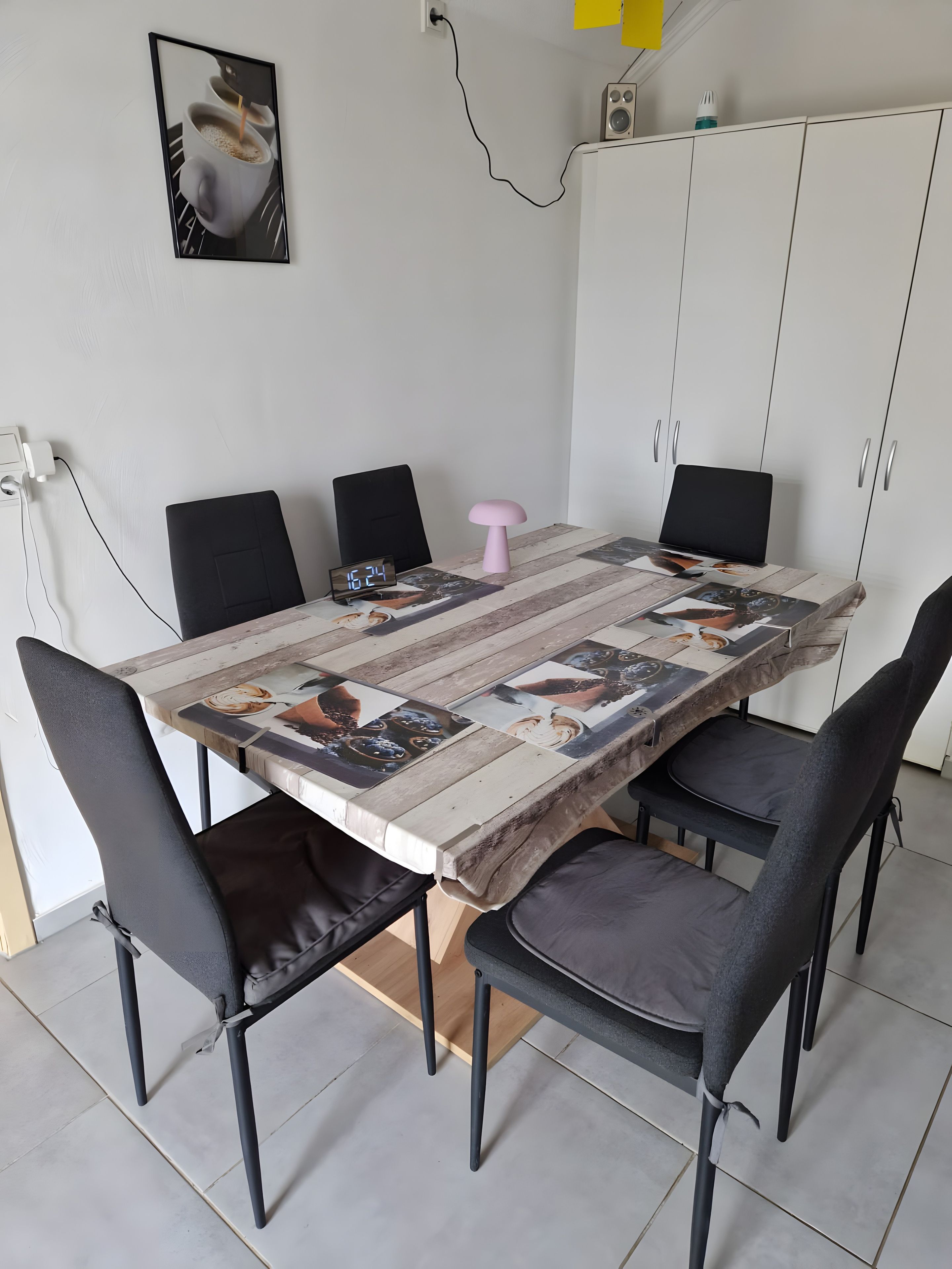 A dining table with a patchwork tablecloth, six chairs, a pink stool, a digital clock, and a speaker on a white cabinet.