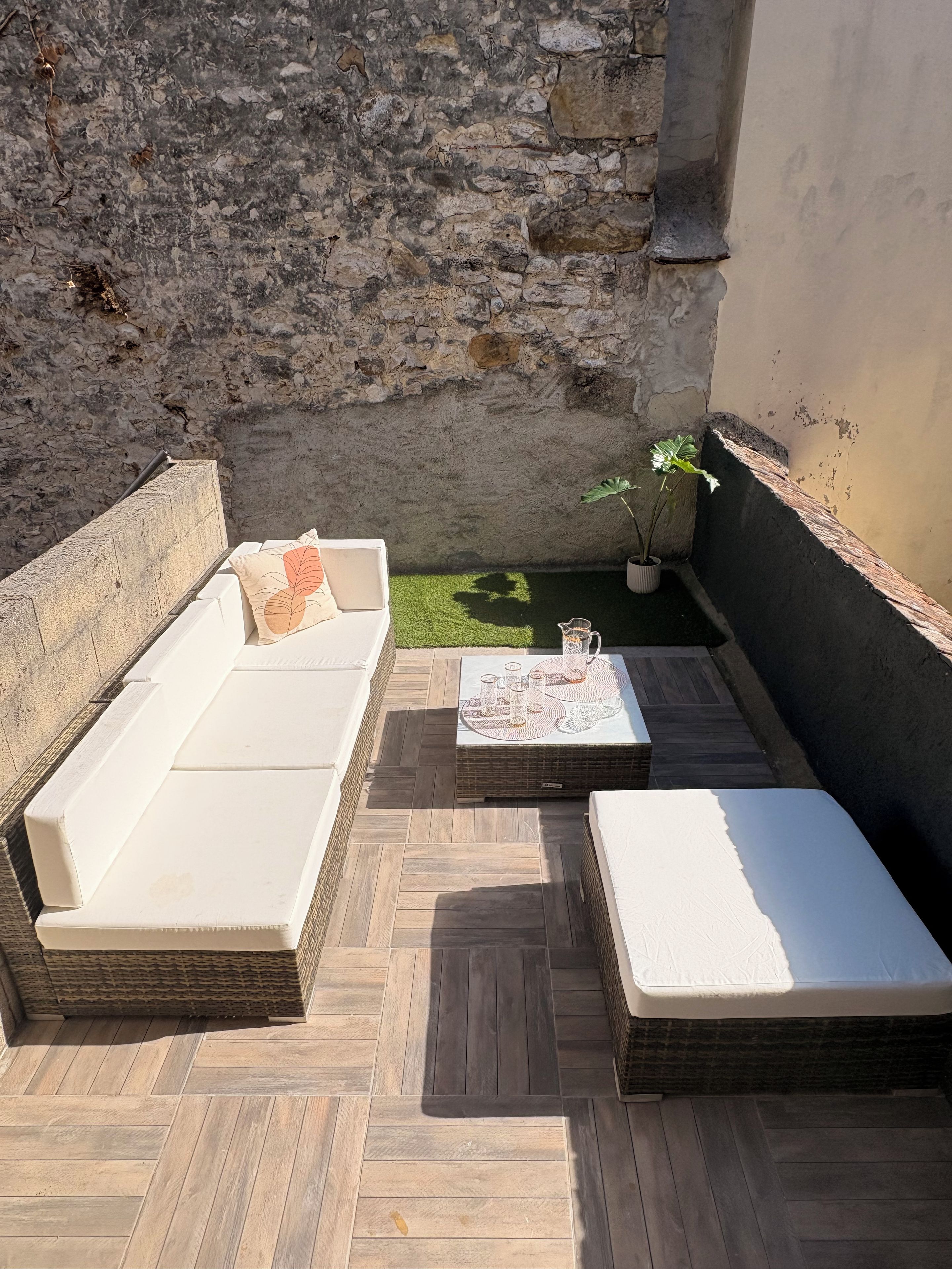 A cozy outdoor seating area with a stone wall, a wooden floor, a white sofa, a small table with drinks, and a potted plant.