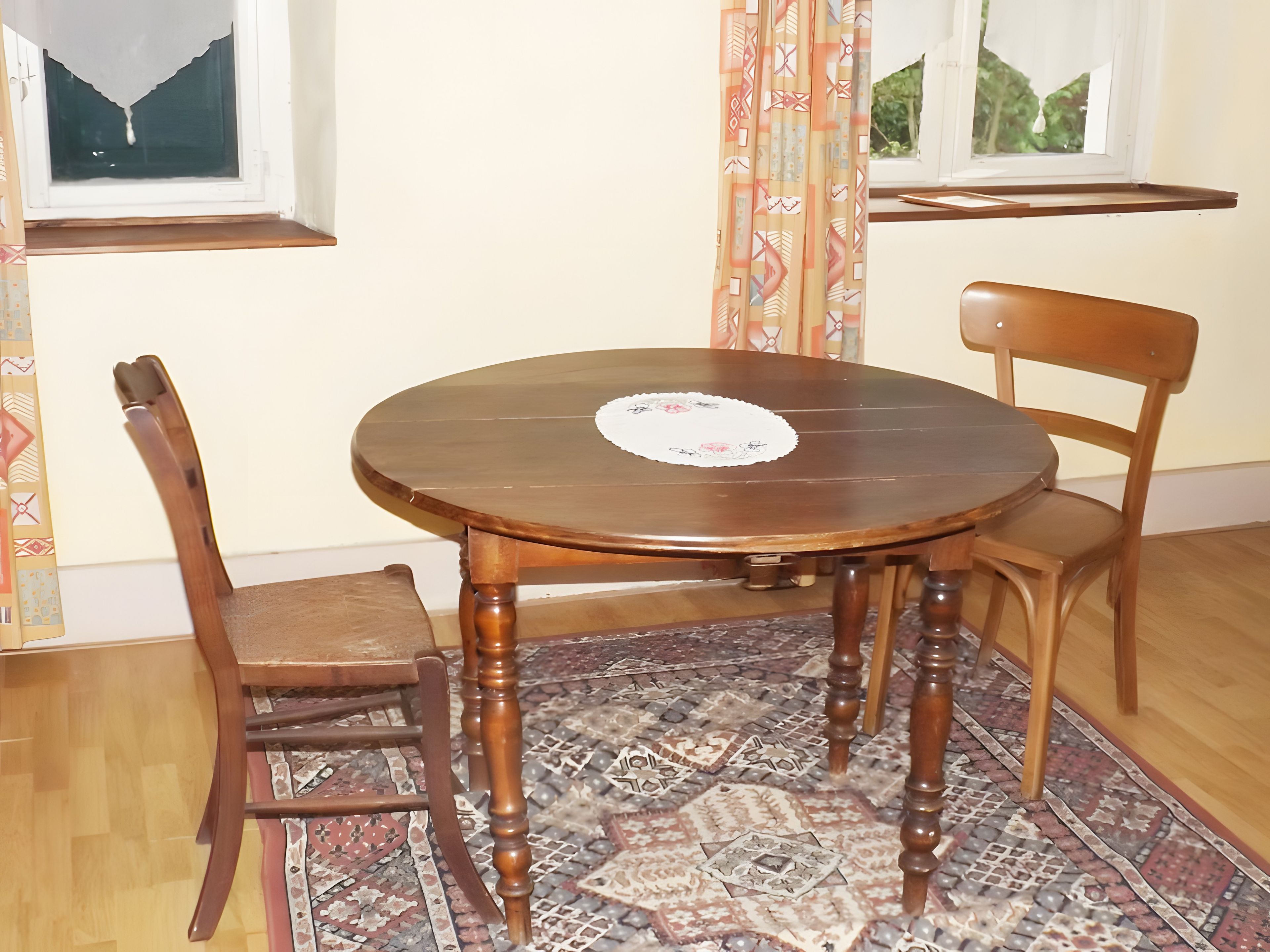 A round wooden dining table with a lace doily, two wooden chairs, a patterned rug, and a window with curtains.