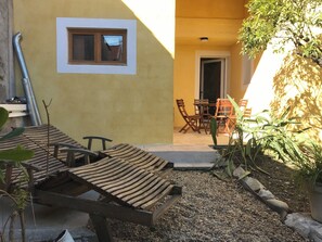 Eine Terrasse mit Holzliegen, einem Tisch und Stühlen, einem Fenster und einer Tür zu einem Innenbereich.