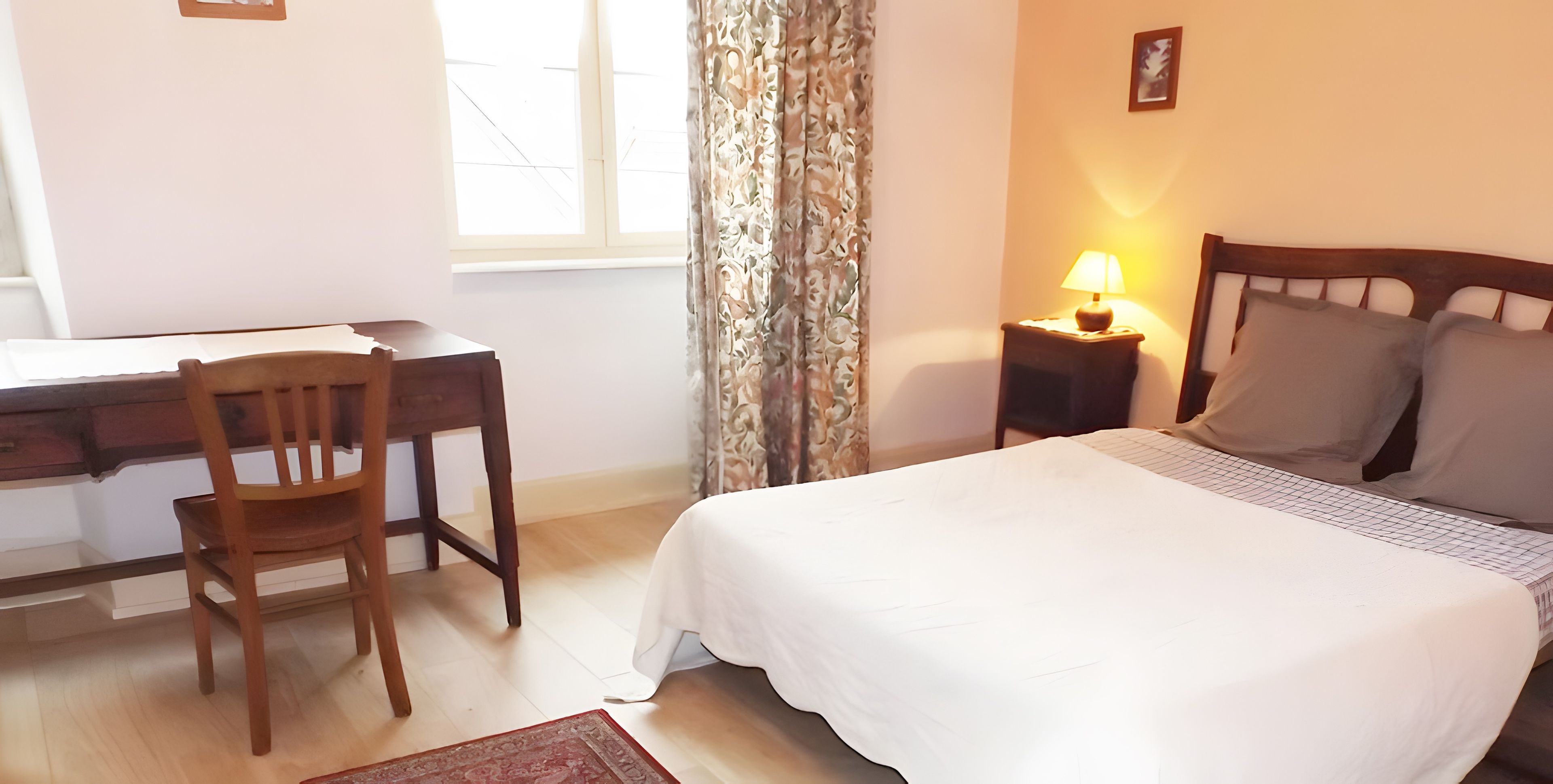A hotel room with a bed, a desk, a chair, a nightstand with a lamp, and a window with curtains.