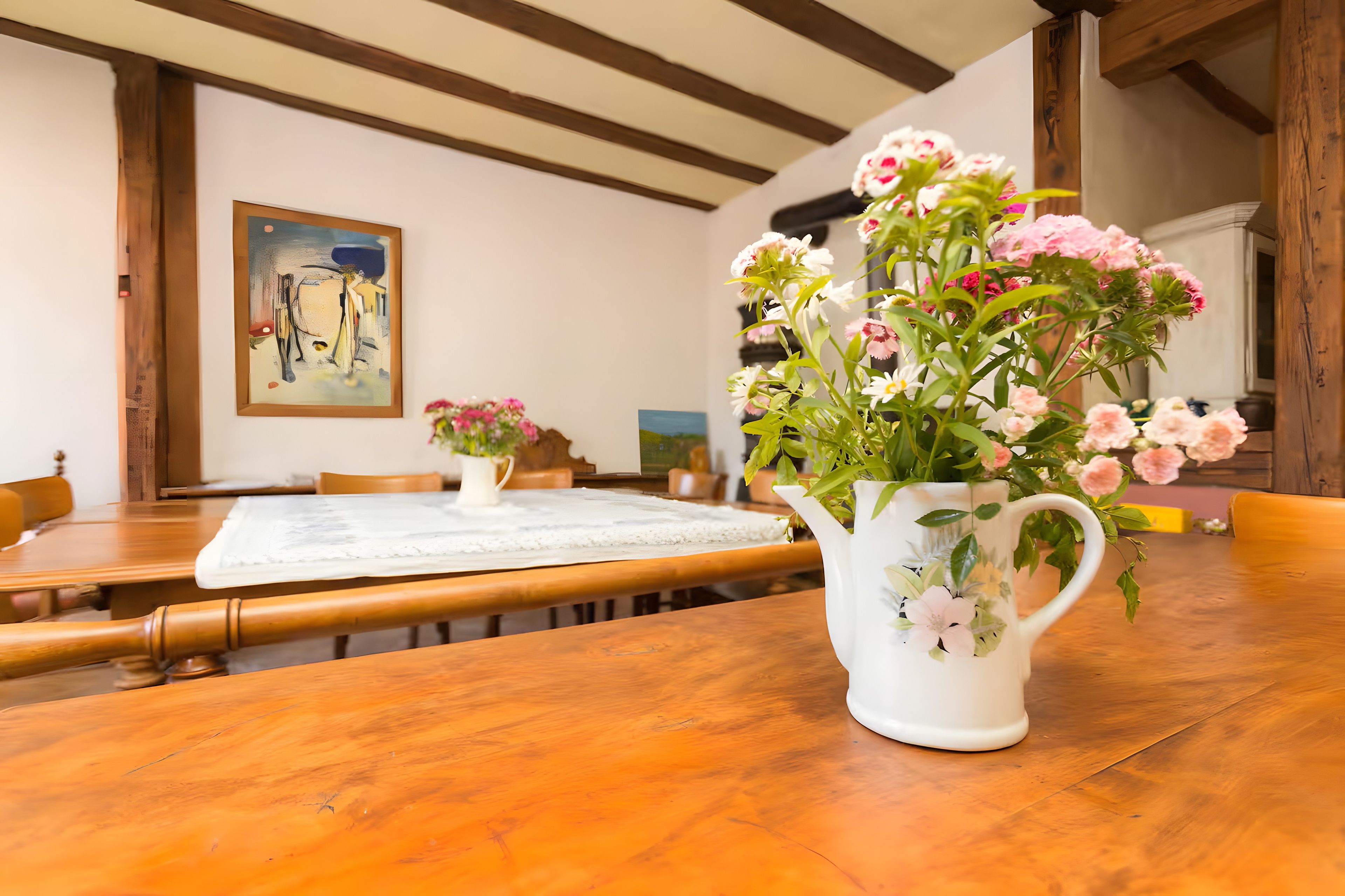 A dining area with a wooden table, chairs, a white pitcher with flowers, and a painting on the wall.