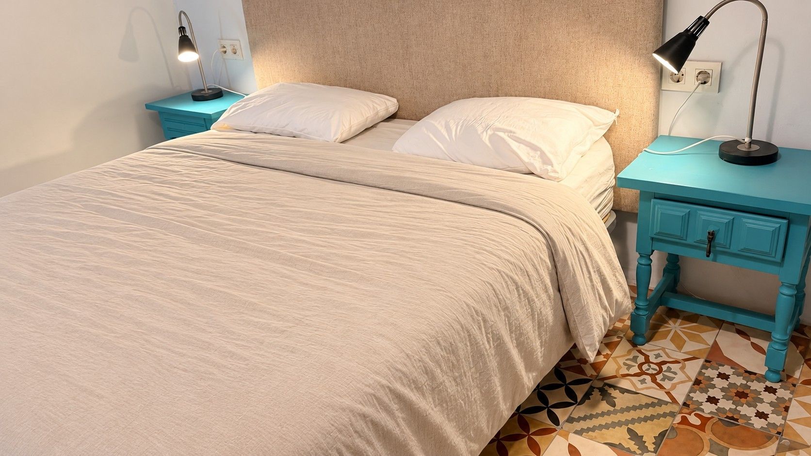 Una cama con una colcha color beige, dos almohadas y un cabecero. Hay dos mesitas de noche con lámparas, una a cada lado de la cama. El piso tiene un diseño decorativo.