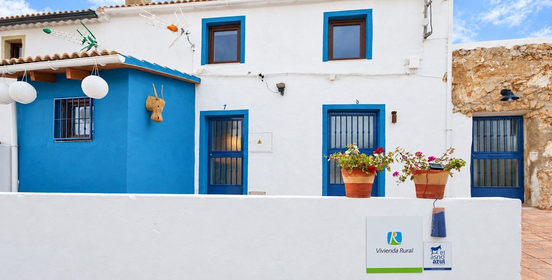Un edificio blanco con puertas y ventanas azules, un letrero con el número 7 y plantas en macetas en el balcón.