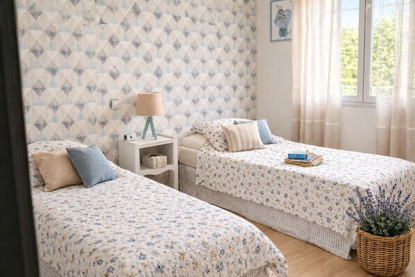 A bedroom with two single beds, a bedside table with a lamp, a small shelf, and a window with curtains. - New Listing (Jonquières)