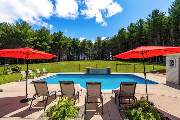A swimming pool with lounge chairs and red umbrellas, surrounded by a grassy area and trees.