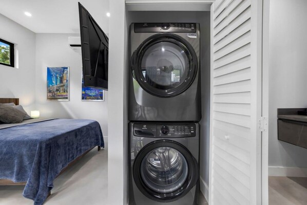 Une petite buanderie équipée d’un lave-linge et d’un sèche-linge à chargement frontal, un lit avec une couverture bleue, un téléviseur fixé au mur et un tableau encadré accroché au mur.