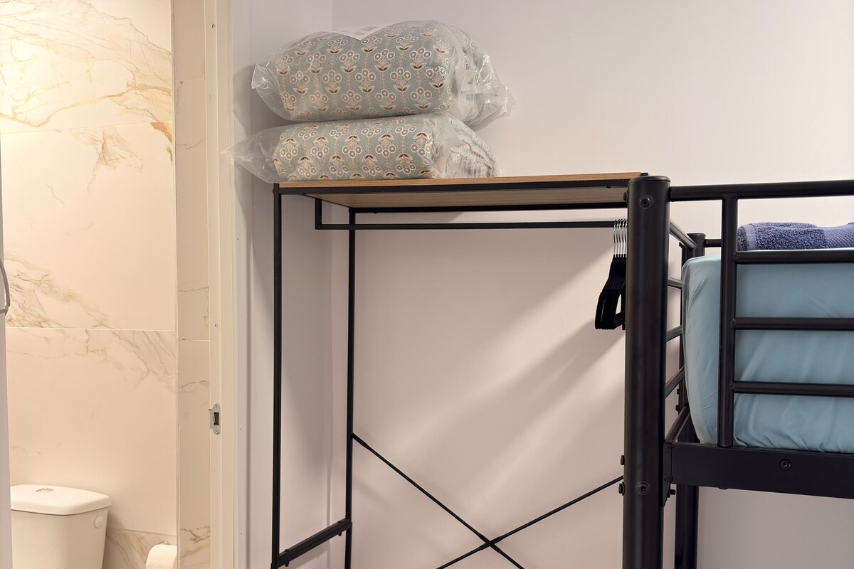 A bunk bed with a control panel on the lower shelf, a small trash bin, and a toilet in the adjacent room.