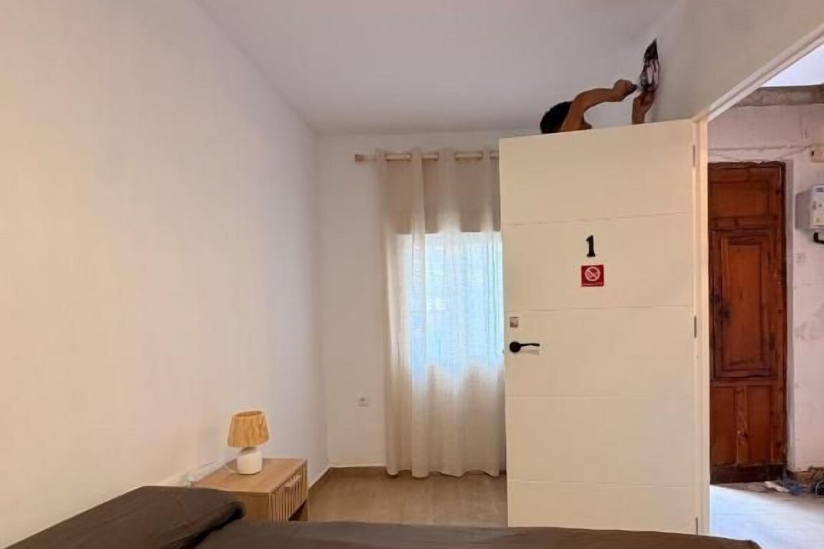 A room with a bed, a nightstand, a lamp, and a door leading to another room.