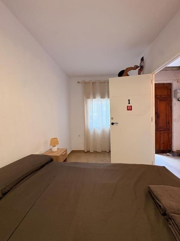 A room with a bed, a nightstand, a lamp, and a door leading to another room. - Cami Real Hostel (Port de Sagunt)
