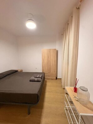 A room with a bed, a wooden wardrobe, a table with a jar and a red object, and a curtain. - Cami Real Hostel (Port de Sagunt)