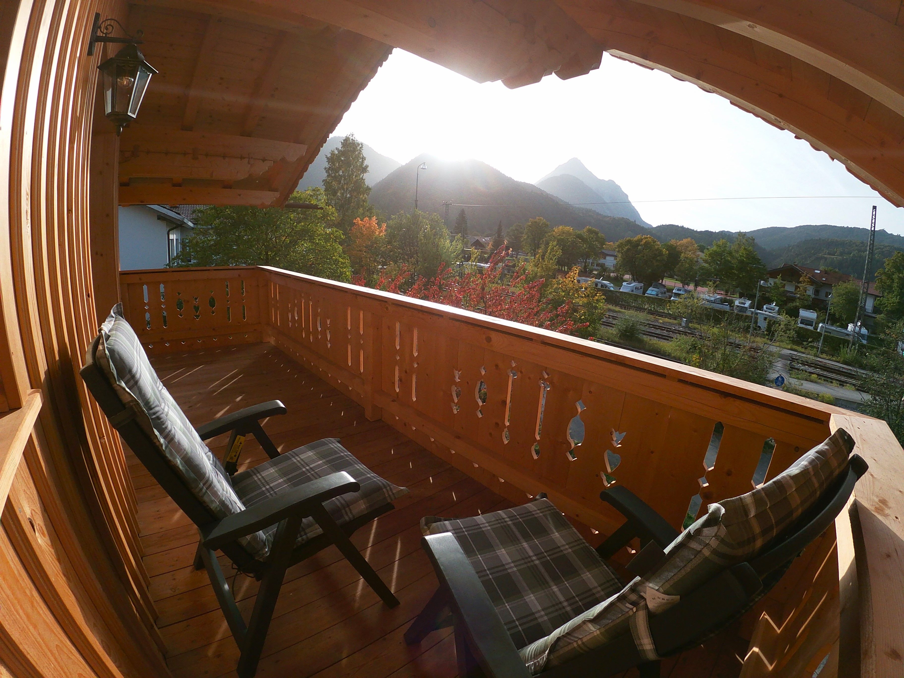 Ein Holzbalkon mit zwei Stühlen und einem Tisch, der auf eine gebirgige Landschaft mit Häusern und Bäumen blickt.