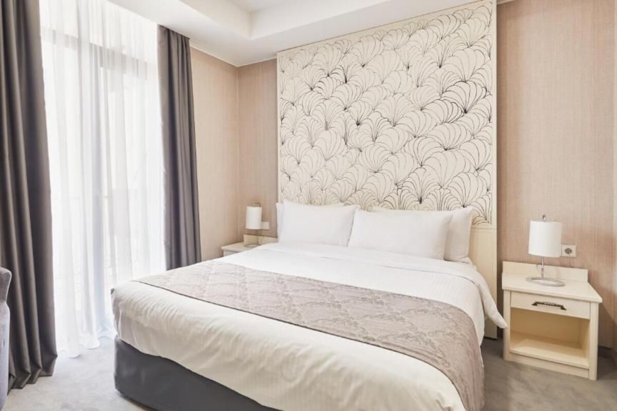 Habitación de hotel con una cama grande, dos mesitas de noche, una silla y un ventanal con cortinas.
