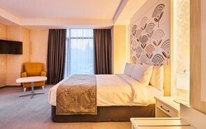 Habitación de hotel con cama, televisor de pantalla plana, mesita, silla y un amplio ventanal con cortinas.
