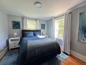 Une chambre à coucher avec un lit, des tables de chevet, une fenêtre avec des rideaux et des œuvres d’art accrochées aux murs.