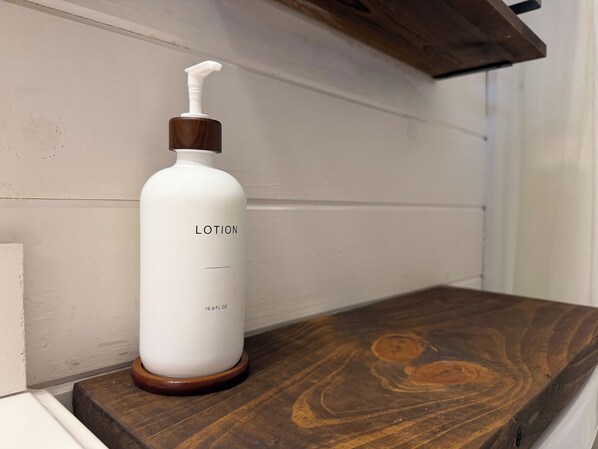 Un flacon de lotion blanche, doté d’un bouchon et d’un pulvérisateur en bois, est posé sur une étagère en bois, adossée à un mur blanc.