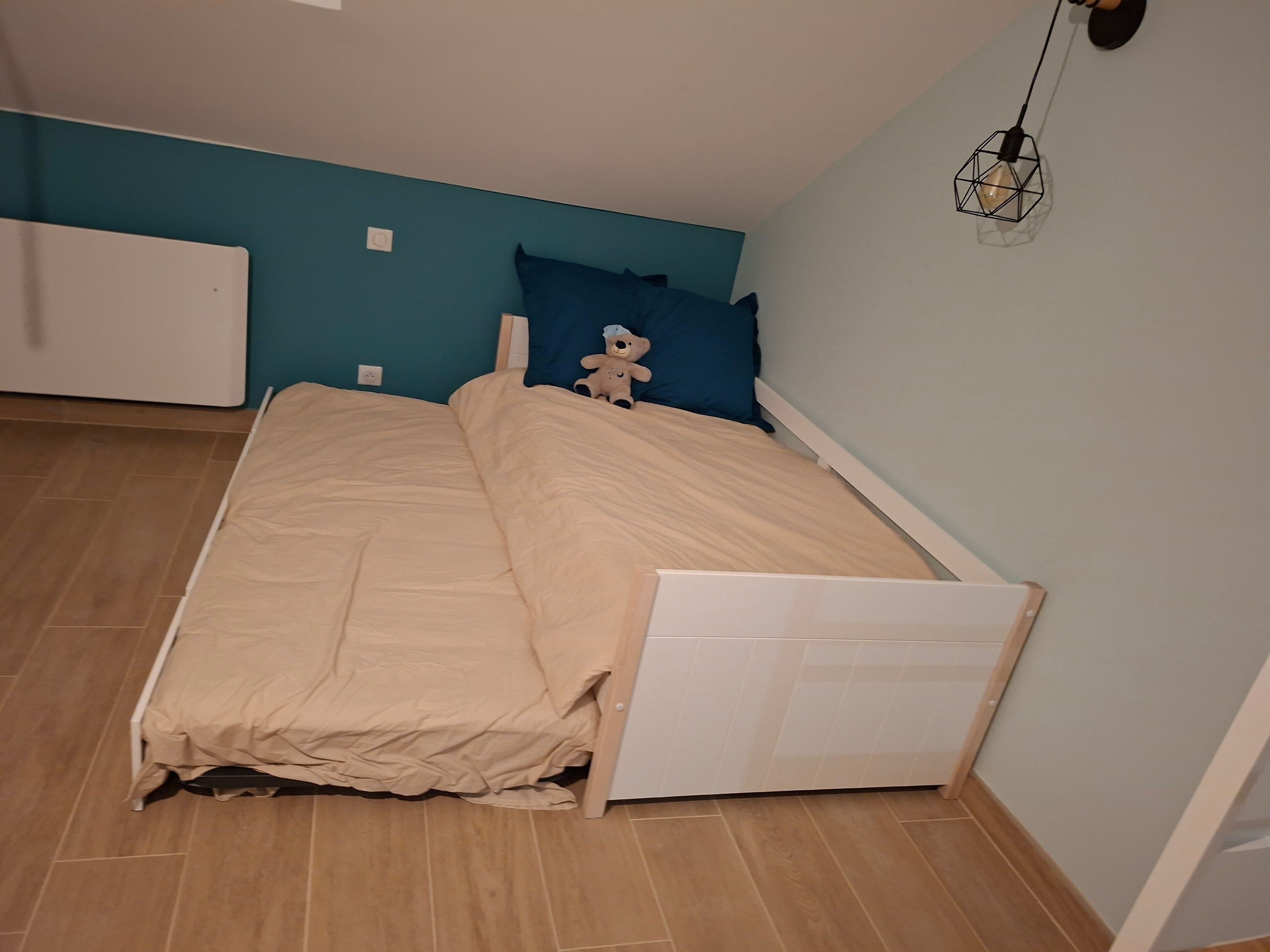 Un lit simple avec un cadre blanc, dans une pièce aux murs de couleur turquoise et au plancher en bois.