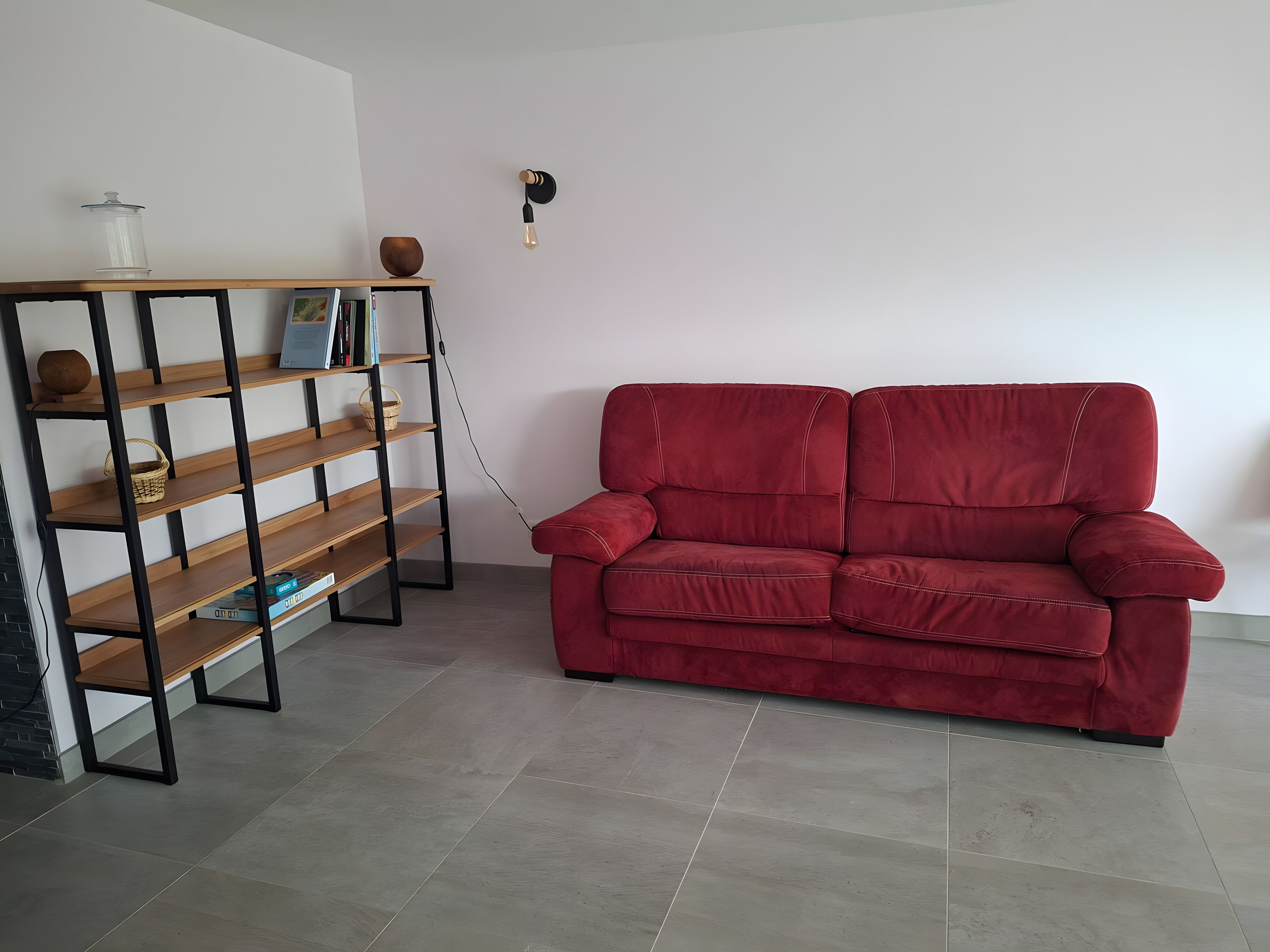 Un canapé rouge, une bibliothèque en bois et un luminaire fixé au mur dans une pièce dont le sol est recouvert de carreaux.