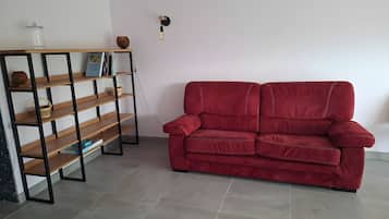 Un canapé rouge, une bibliothèque en bois et un luminaire fixé au mur dans une pièce dont le sol est recouvert de carreaux.