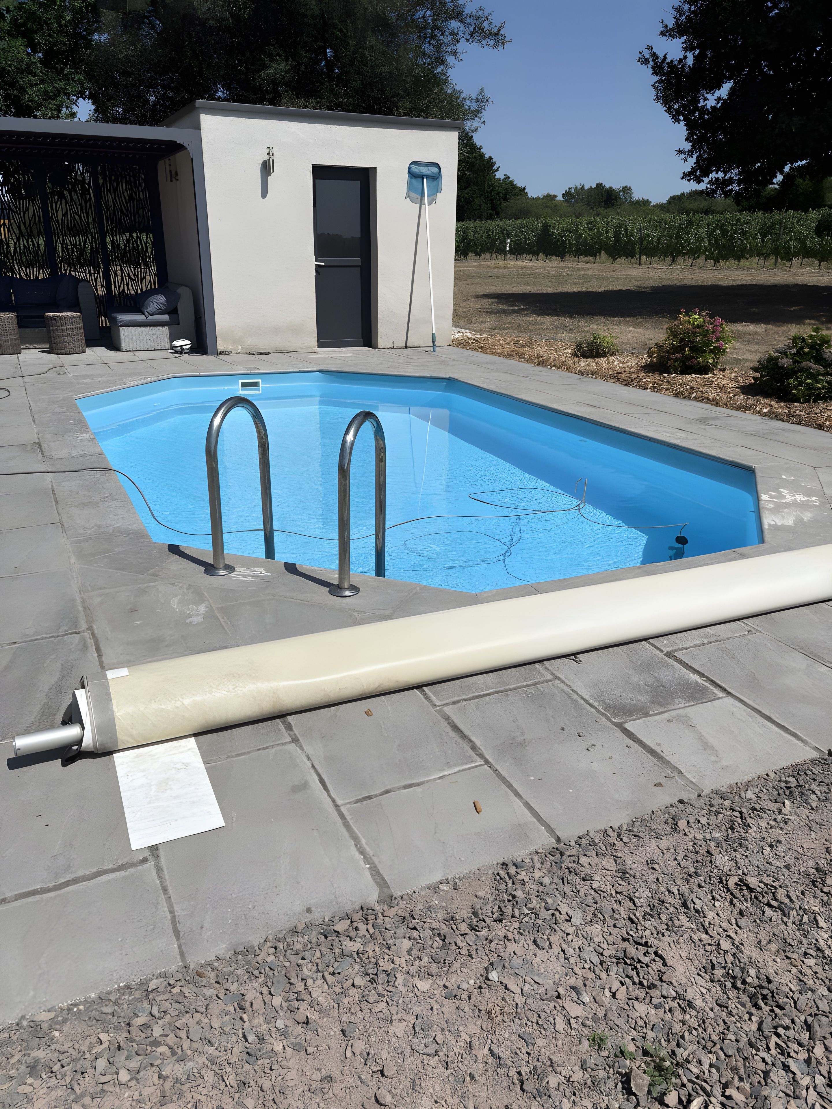 Una piscina rectangular con dos escaleras, un pequeño edificio con una puerta y un jardín con árboles y arbustos.