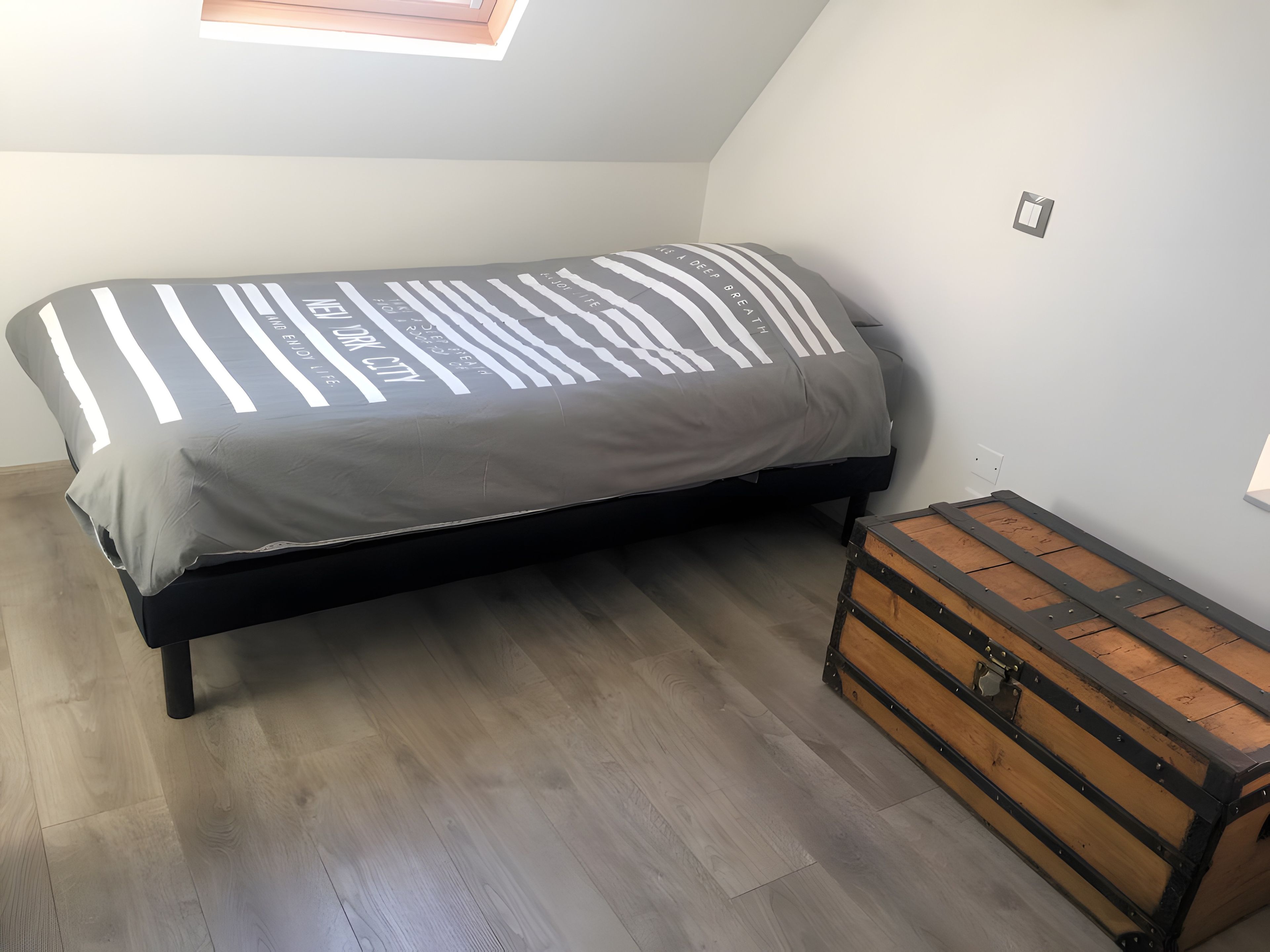 Una cama individual con colcha a rayas, un cofre de madera y un tragaluz en una habitación con suelo de madera.