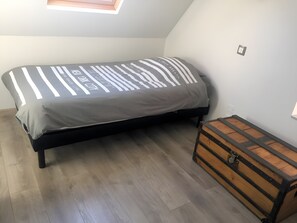 Un lit simple avec une couvre-lit à rayures, une commode en bois et une lucarne dans une pièce dont le sol est recouvert de planches de bois.