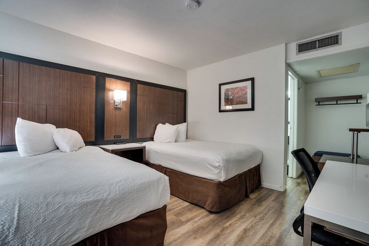 A hotel room with two beds, a desk, a chair, and a dining table.