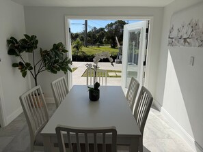 Spiseområde med et bord og stole, en potteplante og udsigt gennem en åben dør til en have.