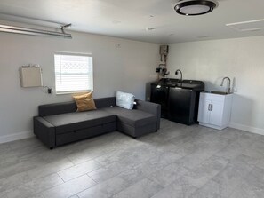 En moderne stue med en hjørnesofa, et indbygget køkkenområde og et vindue med persienner.